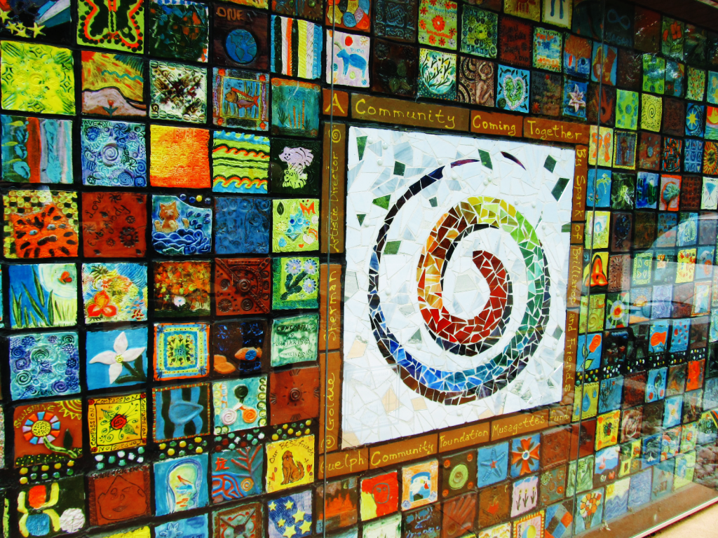 A mosaic wall made up of individual ceramic tiles arranged in a quilt-like pattern. Each tile was individually painted by a member of the community and all are unique. In the center of the mosaic there is a large rainbow swirl done in recycled beach glass. This swirl is the Garden's logo.
